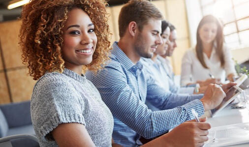 Woman with reddish curly hair sitting with her team at a meeting and smiling into the camera.