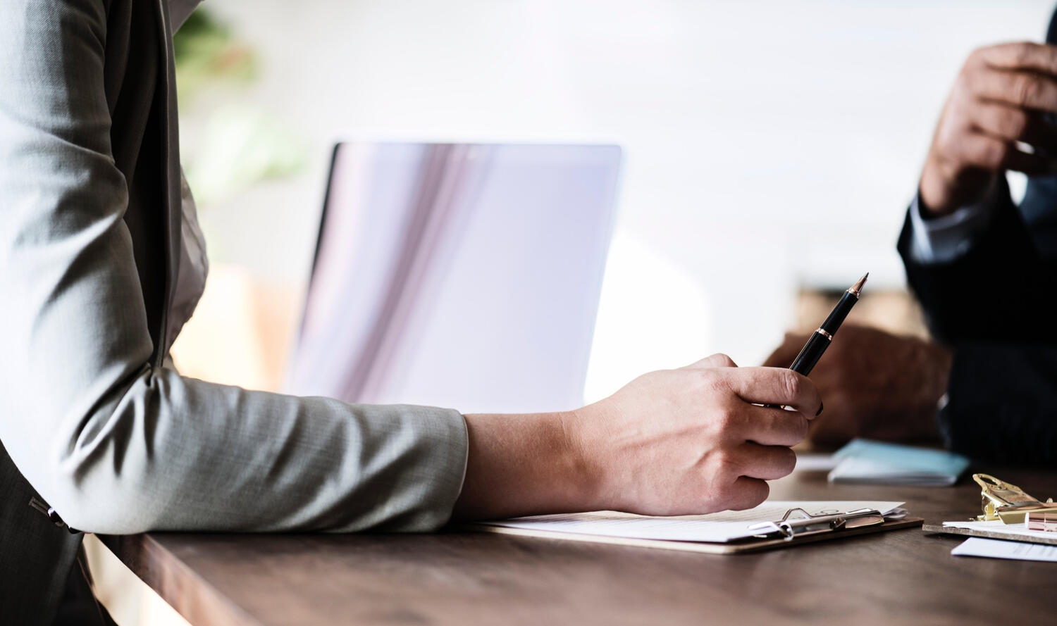 Two individuals sitting at a desk. One of the individuals is taking notes with a paper and pen.